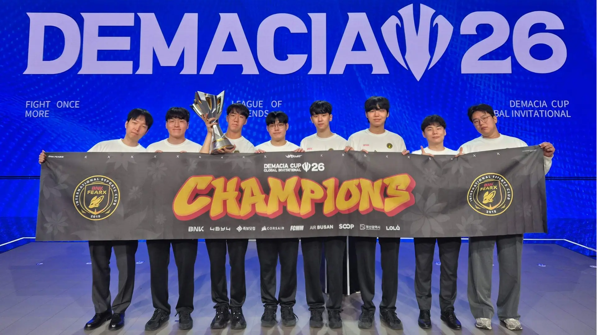 BNK FEARX League of Legends roster posing on stage behind a 'CHAMPIONS' banner while holding the Demacia Cup Global Invitational 2026 trophy, in front of a blue 'DEMACIA 26' event backdrop.