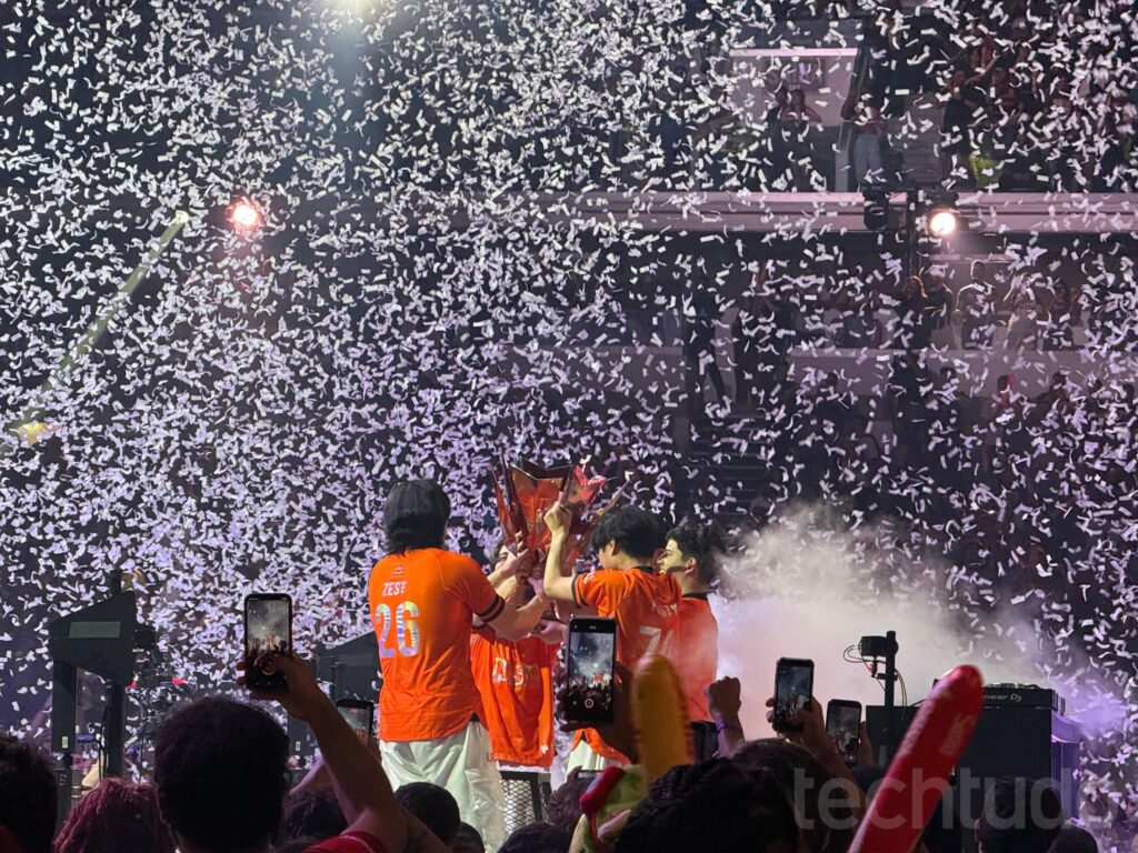 LOS players seen from behind raising the CBLOL trophy under a shower of confetti as fans in the arena record the moment on their phones