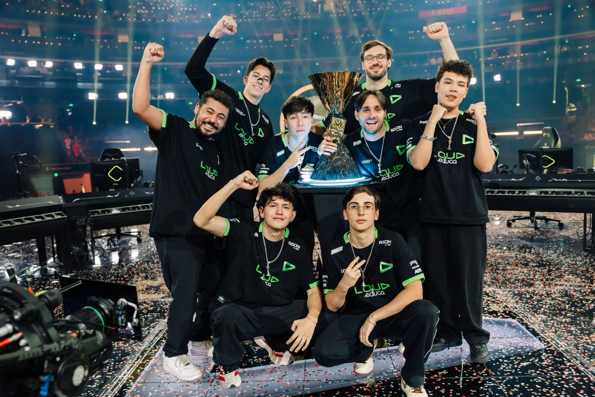 The LOUD VALORANT roster in black-and-green jerseys celebrate on the VALORANT Champions Shanghai stage, fists raised around the Champions trophy with confetti scattered on the floor.