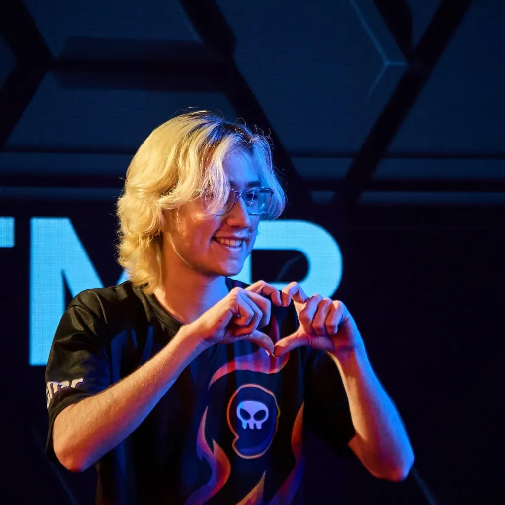 A smiling blonde Overwatch player wearing glasses and a black jersey with a skull logo, making a heart shape with his hands on stage at an OWCS event.