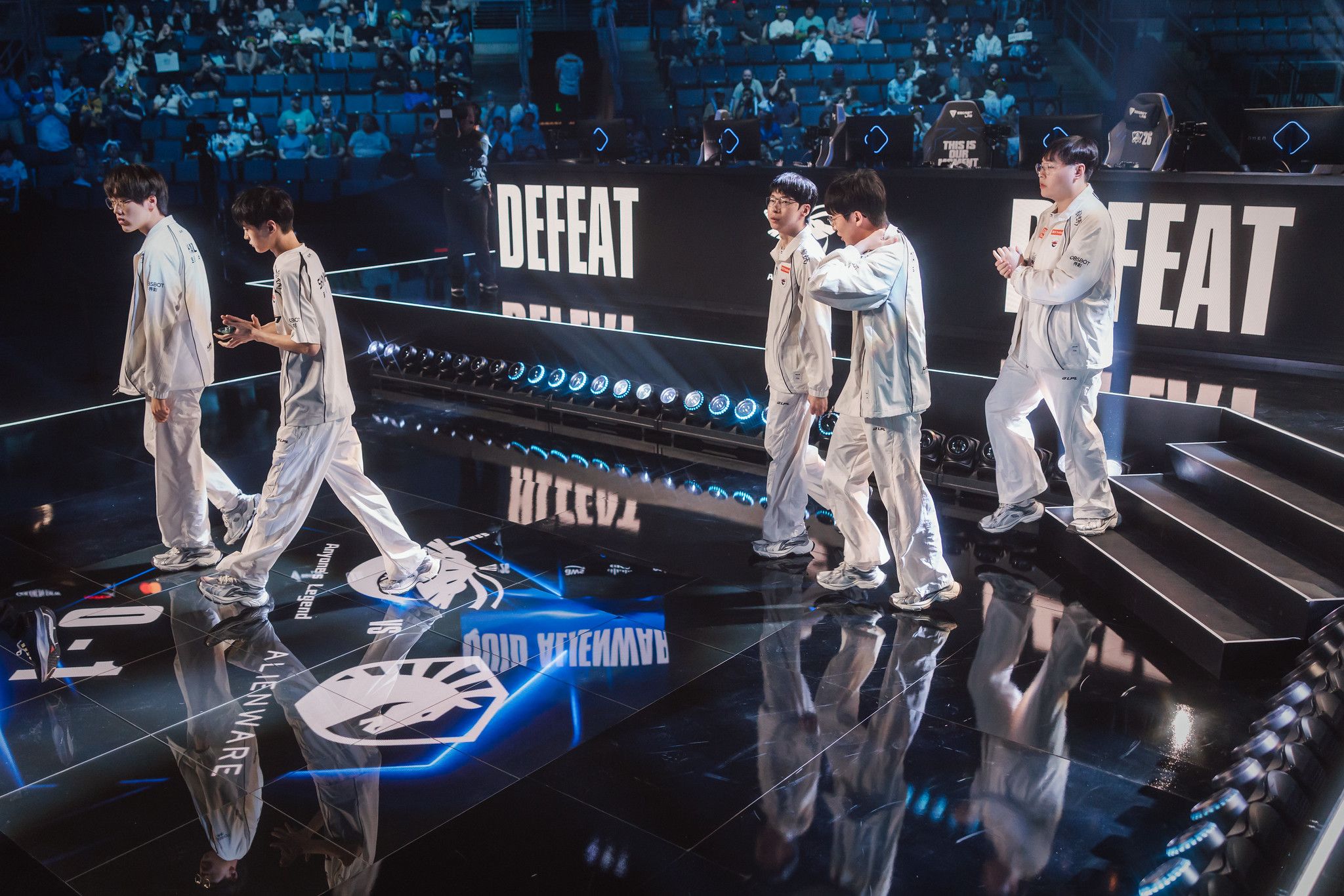 Wide shot of the Worlds 2026 Swiss Stage arena as the Anyone's Legend roster walks off stage beneath large 'DEFEAT' screens, with the Team Liquid and Anyone's Legend logos and a 0-3 score reflected on the glossy stage floor.